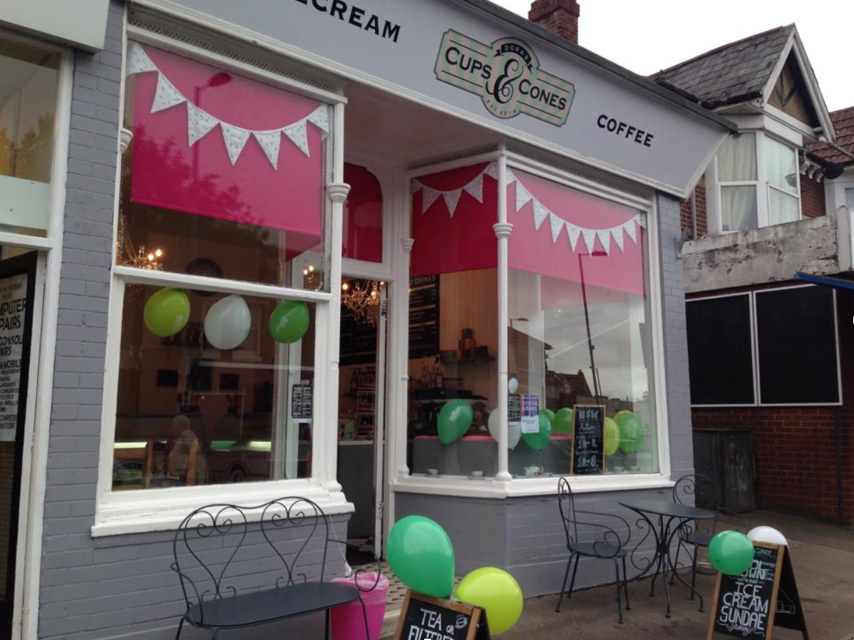 a grey building with white windows and pink blinds called dora's cups & cones ice cream parlour